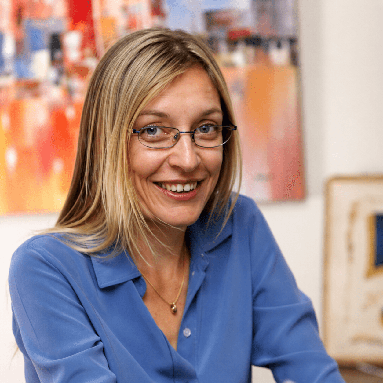Smiling blonde woman wearing glasses and a blue shirt in front of colorful abstract art.