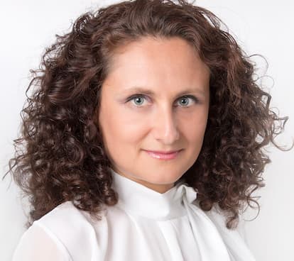 Woman with voluminous curly brown hair and blue eyes wearing a white high-neck blouse.